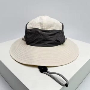 Puede incluir: Sombrero para el sol color crema con ala ancha y corona gris oscuro y crema. El sombrero presenta un logotipo de montaña negro y una correa de barbilla ajustable. El sombrero está diseñado para actividades al aire libre.