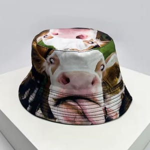Puede incluir: Un sombrero de pescador con una imagen en primer plano de la cara de una vaca. El sombrero tiene un diseño de cara de vaca blanca y marrón con la lengua de la vaca saliendo. El sombrero está sobre una superficie blanca.