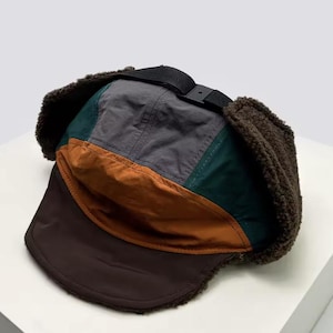 Puede incluir: Un gorro de invierno multicolor con orejeras. El gorro presenta una combinación de colores marrón, gris, verde y naranja. Las orejeras son de un material marrón difuso. Una correa negra con hebilla está en la parte superior.