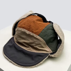 Peut inclure: Un chapeau de trappeur multicolore avec un bord noir et des cache-oreilles. Le chapeau présente des panneaux de tissu marron, vert olive et beige, avec une doublure douce et duveteuse autour des oreilles. Une sangle noire est visible à l'arrière.