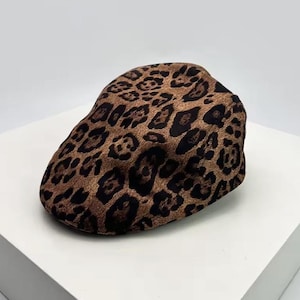 Peut inclure: Casquette de style gavroche avec un imprimé léopard marron et noir. La casquette a une forme arrondie classique et est faite d'une matière douce. Le motif léopard présente des taches marron foncé sur un fond marron clair.