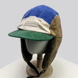 Puede incluir: Gorra de cazador de pana en azul, beige, verde y marrón. La gorra tiene una visera corta verde, orejeras y un pequeño parche rectangular con el texto "Ludespac". La gorra se exhibe en una cabeza de maniquí.