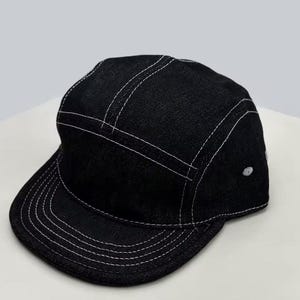 May include: Black denim cap with a flat peak and a five-panel design. The cap has white stitching along the seams and peak, and a small white button on the side. A stylish accessory for casual wear.