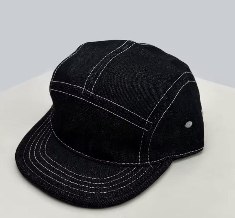 May include: Black denim cap with a flat peak and a five-panel design. The cap has white stitching along the seams and peak, and a small white button on the side. A stylish accessory for casual wear.