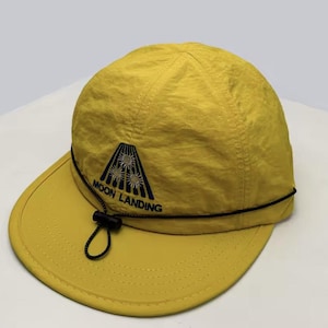 Peut inclure: Casquette en nylon jaune avec une visière incurvée et un cordon de serrage réglable. Le chapeau présente un graphisme noir d'une fusée stylisée et les mots "MOON LANDING" en noir. La casquette est de couleur jaune uni.