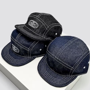 May include: Three denim five-panel caps in shades of blue and black. Each cap has white stitching and a small, rectangular patch with text. The caps have a flat peak and a classic, casual style.