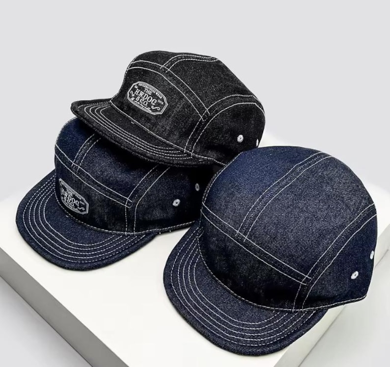 May include: Three denim five-panel caps in shades of blue and black. Each cap has white stitching and a small, rectangular patch with text. The caps have a flat peak and a classic, casual style.