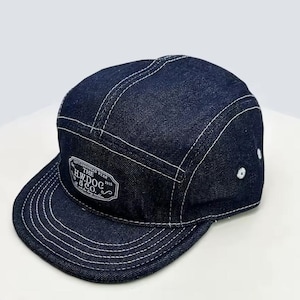 May include: A dark blue denim cap with a flat peak and white stitching. The cap features a white patch with the text "THE H.W.DOG & CO. 1916". The cap has a five-panel design.