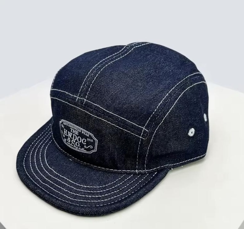 May include: A dark blue denim cap with a flat peak and white stitching. The cap features a white patch with the text "THE H.W.DOG & CO. 1916". The cap has a five-panel design.