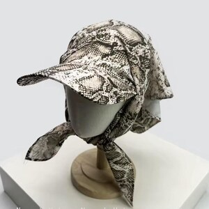 May include: A snakeskin print hat with a visor and attached scarf. The hat is displayed on a mannequin head. The print features shades of black, brown, and cream. The scarf is tied in a bow under the chin.