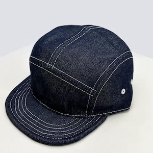 Könnte beinhalten: Dunkelblaue Jeans-Cap mit flachem Schirm. Die Cap hat weiße Nähte entlang der Nähte und des Schirms sowie weiße Ösen an der Seite. Die Cap ist im Fünf-Panel-Stil gehalten.