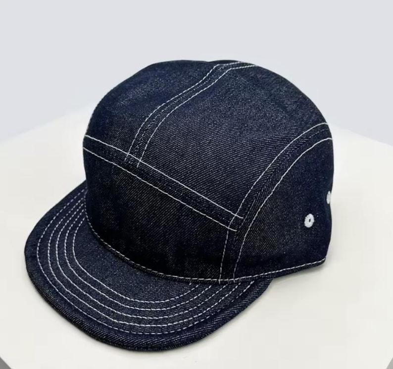 May include: A dark blue denim cap with a flat peak. The cap has white stitching along the seams and peak, and white eyelets on the side. The cap is a five-panel style.