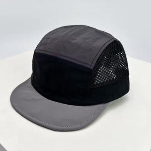 Puede incluir: Gorra de cinco paneles gris y negra con visera curva. La gorra presenta un panel de malla negra en el lateral para ventilación. La visera y el panel frontal son grises, mientras que el panel central es negro.