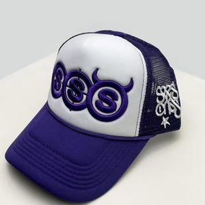 Peut inclure: Casquette de camionneur violette et blanche avec une visière incurvée. Le panneau avant présente un motif brodé violet avec les lettres "SSS". L'arrière en mesh est bleu marine et la casquette a une fermeture à pression.