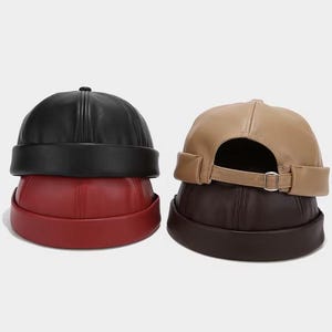 Op de afbeelding: Drie leren dockerhoeden in zwart, bordeauxrood en bruin. De hoeden hebben een ronde kroon en een korte rand. De bruine hoed heeft een beige band met een zilveren gesp. De hoeden zijn op elkaar gestapeld.