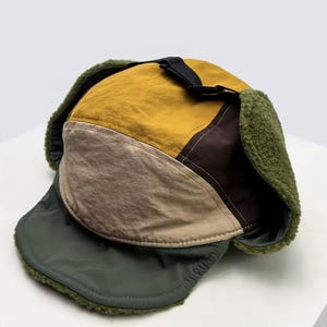 Peut inclure: Un chapeau de trappeur multicolore en jaune moutarde, vert olive, marron et beige. Le chapeau est doté d'une sangle noire et d'une doublure douce et duveteuse autour des cache-oreilles et du bord. Un accessoire d'hiver élégant et protecteur.