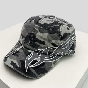 Peut inclure: Casquette de baseball camouflage grise et noire. La casquette a une visière incurvée avec un motif de flamme blanche. Le chapeau est en tissu doux et a une couronne arrondie. Un accessoire de mode.