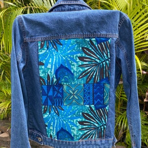 May include: A blue denim jacket with a colorful patterned fabric patch on the back. The patch features a tropical leaf design in shades of blue, green, and black.