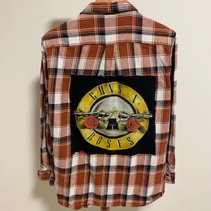 May include: A brown, white, and black plaid flannel shirt with a Guns N' Roses patch on the back. The patch features a yellow emblem with a gun, roses, and the band's name.