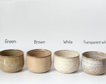 May include: Four ceramic bowls in varying colors: green, brown, white, and transparent white. Each bowl has a rounded shape and a speckled texture. The bowls are arranged in a row against a white background.