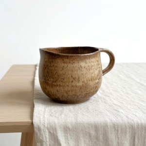 Puede incluir: Una taza de cerámica marrón hecha a mano, con un cuerpo redondeado y un asa pequeña. La taza tiene una superficie texturizada y un borde marrón oscuro. Se encuentra sobre un mantel de lino claro, con una mesa de madera en el fondo.