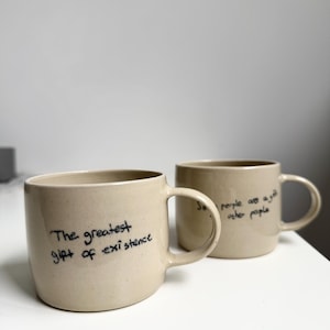 Könnte beinhalten: Zwei beige Keramikbecher mit gesprenkelter Textur und abgerundeten Henkeln. Der vordere Becher trägt den schwarzen handschriftlichen Text "The greatest gift of existence", der hintere Becher zeigt "Some people are a gift to other people."