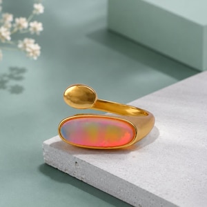 May include: A gold-toned open ring featuring a smooth, polished band that terminates in a small circular accent on one end and a larger, horizontally oriented oval setting on the other, which contains a smooth, iridescent, multi-colored stone.