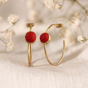 May include: A pair of gold-toned hoop earrings, each featuring a single, smooth, circular red stone set in a bezel. The earrings have a thin, polished metal wire frame that forms a circular shape, with the red stone positioned on the upper curve.