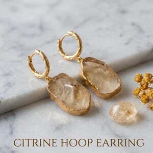 Può includere: Orecchini a cerchio dorati con pietre di citrino. Gli orecchini hanno un design a cerchio strutturato e presentano pietre di citrino giallo chiaro di forma irregolare con bordi dorati. L'immagine include anche una pietra di citrino sciolta e il testo "CITRINE HOOP EARRING".