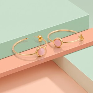 Puede incluir: Un par de pendientes de aro dorados, cada uno con una piedra redonda, lisa y de color rosa claro engastada en el centro del fino alambre de metal. Los pendientes tienen un diseño sencillo de aro abierto con un pequeño cierre de perno.