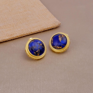 May include: A pair of round stud earrings featuring a deep blue stone with gold-coloured veining, set within a textured gold-tone metal frame. The circular design highlights a polished finish on the stones and a beaded edge on the metal setting.