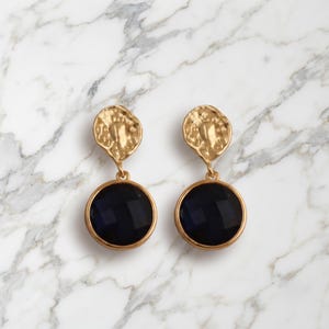 Peut inclure: Une paire de boucles d'oreilles pendantes dorées composées d'une pièce supérieure circulaire à la texture irrégulière, reliée à une pierre ronde facettée d'un bleu marine profond. Les boucles présentent une finition métallique polie.