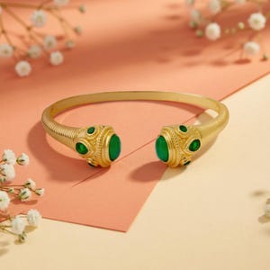 Könnte beinhalten: Ein goldfarbenes, offenes Armband mit einem strukturierten, gerippten Band, das in zwei verzierten, abgerundeten Enden endet. Jedes Ende ist mit einem großen, glatten, ovalen grünen Stein verziert, der von kleineren, tropfenförmigen grünen Akzenten umgeben ist.