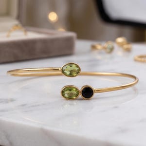 Op de afbeelding: Een goudkleurige open armband met drie gefacetteerde edelstenen. Het ene uiteinde bevat een ovaal geslepen lichtgroene peridoot, terwijl het andere uiteinde een bijpassende ovale lichtgroene peridoot naast een ronde, zwarte onyxsteen toont.
