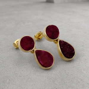 May include: A pair of gold-toned stud earrings featuring a dual-stone design. Each earring consists of a circular deep magenta stone positioned above a larger, teardrop-shaped stone of the same dark pink color, all set in polished gold metal.