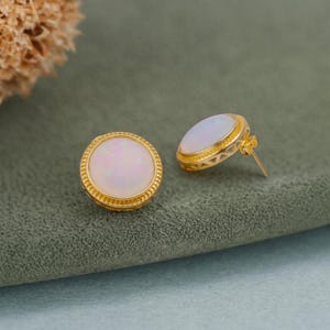 May include: A pair of round stud earrings featuring smooth, iridescent white opal cabochons set in gold-toned metal. The circular settings have a textured, beaded edge detail, and the earring posts are visible on the back of one piece.