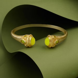 Puede incluir: Un brazalete abierto de tono dorado con una textura acanalada a lo largo de la banda y dos extremos decorativos. Cada extremo está adornado con una piedra grande, lisa y ovalada de color amarillo brillante, flanqueada por piedras más pequeñas.