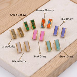 Puede incluir: Una colección de pendientes rectangulares con diversas piedras engastadas en metal dorado. El conjunto incluye Labradorita, Mohave verde, Mohave naranja, Druzy azul, Druzy blanco, Druzy rosa y Druzy verde, cada uno etiquetado con su nombre.