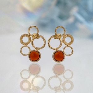 Peut inclure: Une paire de boucles d'oreilles dorées composées d'un groupe abstrait de cercles interconnectés. Chaque boucle présente une pierre ronde et lisse de couleur orange brûlé à la base, avec un mélange d'anneaux en métal poli et texturé.