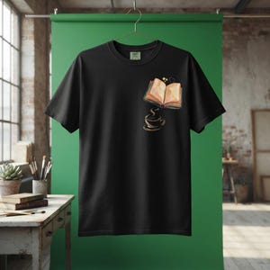 Può includere: Una t-shirt nera a maniche corte con una grafica sulla parte superiore sinistra del petto. Il disegno raffigura un libro aperto con pagine marrone chiaro, una piccola tazza di caffè scuro sotto e due piccole stelle gialle e un cuore sopra.