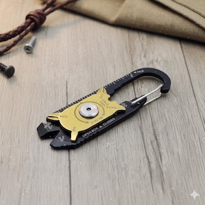 Könnte beinhalten: Ein schwarzes und goldenes Multi-Tool mit einem Karabinerhaken. Das Werkzeug verfügt über ein Lineal, eine Nagelfeile, einen Drahtabisolierer und ein rotierendes Mittelteil. Das Werkzeug ist für verschiedene Aufgaben konzipiert und hat ein kompaktes, tragbares Design.