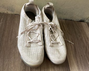 May include: Pair of off-white athletic shoes with woven uppers and matching laces. The shoes have a low-profile design with a clear sole. The shoes are displayed on a wooden surface.