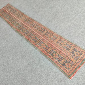 Könnte beinhalten: Ein rechteckiger Läufer mit einem zentralen Design in Pfirsich-, Grau- und Orangetönen. Der Teppich hat ein sich wiederholendes geometrisches Muster und einen roten Rand. Der Teppich liegt auf einer grauen Oberfläche.