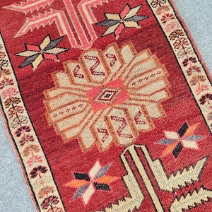 May include: A rectangular runner rug featuring a deep red background with a large central beige and pink geometric floral medallion. The rug is decorated with smaller star-shaped motifs and has a border with repeating floral patterns in beige, pink, and black.