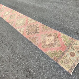 May include: A long, rectangular runner rug with a distressed, vintage appearance. The rug features a repeating geometric pattern in shades of pink, brown, green, and beige. The rug is placed on a dark grey asphalt surface.