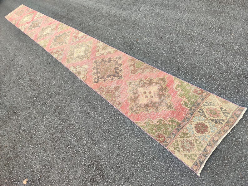 May include: A long, rectangular runner rug with a distressed, vintage appearance. The rug features a repeating geometric pattern in shades of pink, brown, green, and beige. The rug is placed on a dark grey asphalt surface.