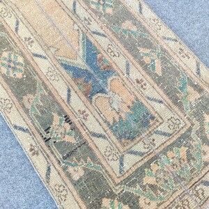 May include: A rectangular, distressed rug with a faded, vintage aesthetic. The rug features a central design with a beige background, bordered by olive green and beige stripes. Floral and geometric patterns in blue, beige, and teal add detail. Frayed edges complete the look.