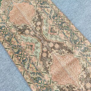 May include: A distressed, rectangular rug with a vintage look. The rug has a central design in peach and brown, framed by a border of teal and green floral patterns. The rug's design is symmetrical and detailed, with a faded appearance.