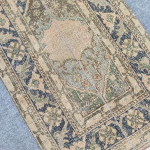 May include: A rectangular rug with an intricate pattern. The rug features a central beige field with a floral design, framed by a border of navy blue and green floral motifs. The rug has a distressed, vintage appearance.