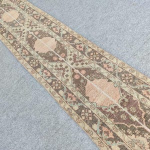Peut inclure: Un tapis rectangulaire à motifs avec un motif central de formes marron clair et pêche, bordé de motifs floraux et géométriques complexes dans des tons de marron, vert et pêche.